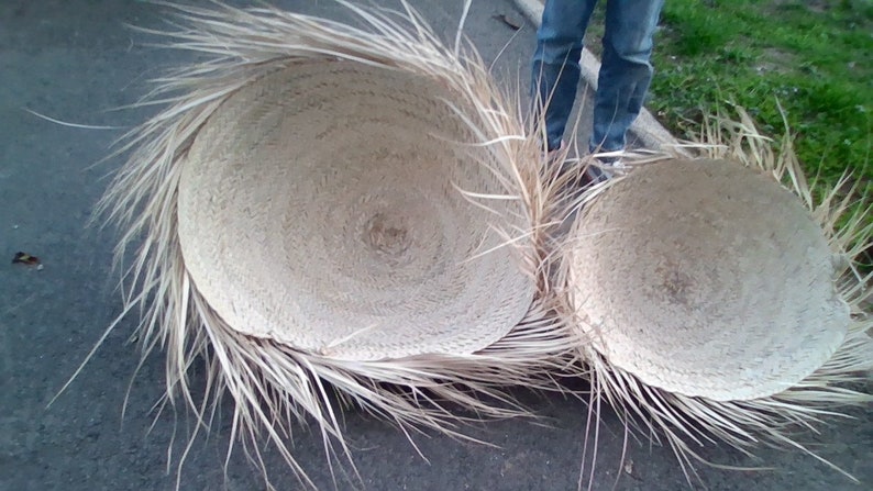 Op de afbeelding: Twee grote, ronde, geweven manden met een natuurlijke, lichtbruine kleur. De manden zijn gemaakt van een natuurlijk materiaal, mogelijk palmbladeren, en hebben een gefranst randje. De manden staan op een grijs oppervlak.
