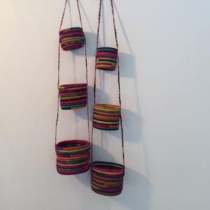May include: Five colorful hanging planters made of woven strips of fabric. The planters are suspended from the ceiling by strings and are arranged in a staggered pattern.