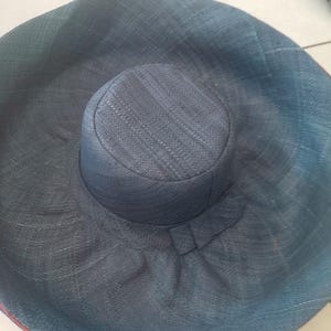 Puede incluir: Un sombrero de sol grande y de ala ancha hecho de paja tejida. El sombrero es de color azul oscuro y tiene una corona ligeramente redondeada.