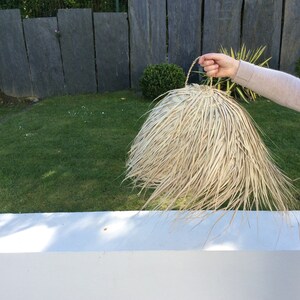 Op de afbeelding: Een grote, ronde, natuurlijke geweven lampenkap met een handvat. De lampenkap is gemaakt van lichtbruin, stroomachtig materiaal en heeft een gefranst randje.