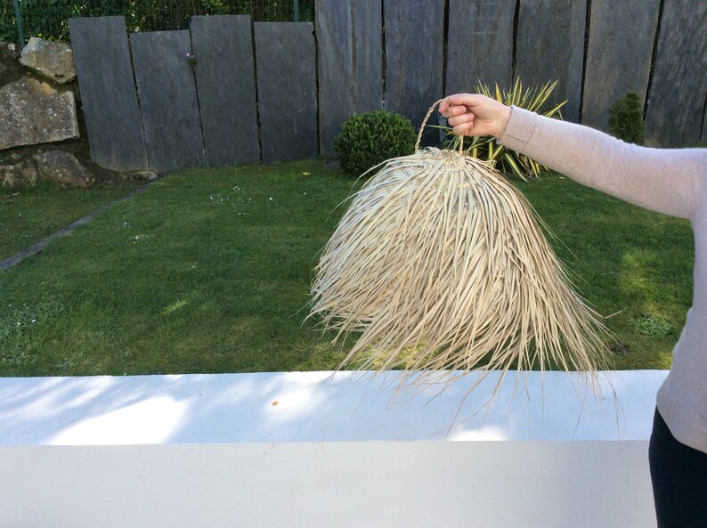 Op de afbeelding: Een grote, ronde, natuurlijke geweven lampenkap met een handvat. De lampenkap is gemaakt van lichtbruin, stroomachtig materiaal en heeft een gefranst randje.