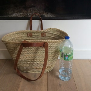May include: A large, oval-shaped straw basket with brown leather handles. The basket is sitting on a wooden floor next to a clear plastic water bottle.