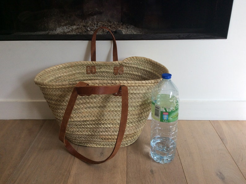 May include: A large, oval-shaped straw basket with brown leather handles. The basket is sitting on a wooden floor next to a clear plastic water bottle.