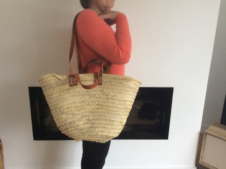 Puede incluir: Un gran bolso de mano de paja natural tejido con asas y correas de cuero marr&oacute;n. El bolso lo lleva una persona que lleva un jersey coral y pantalones negros.
