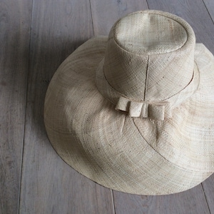 Puede incluir: Un sombrero de paja de ala ancha con un ala grande y flexible y un simple detalle de lazo. El sombrero est&aacute; hecho de un material natural tejido y tiene un color beige claro.