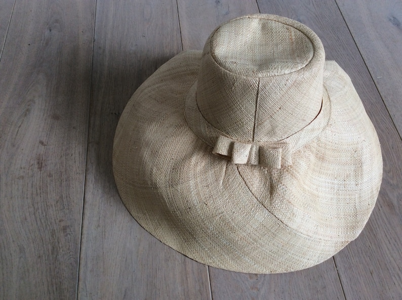 Puede incluir: Un sombrero de paja de ala ancha con un ala grande y flexible y un simple detalle de lazo. El sombrero est&aacute; hecho de un material natural tejido y tiene un color beige claro.