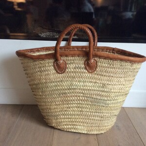 May include: A large, woven straw tote bag with brown leather handles and trim. The bag is empty and sitting on a wooden floor.