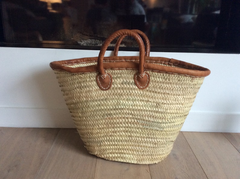 May include: A large, woven straw tote bag with brown leather handles and trim. The bag is empty and sitting on a wooden floor.