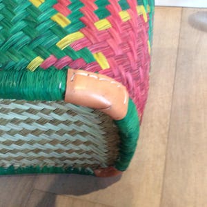 May include: Close-up of a woven basket with a green, red, and yellow pattern. The basket has a brown leather trim and a woven straw base.