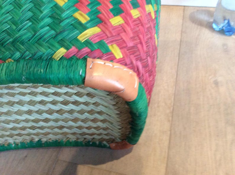 May include: Close-up of a woven basket with a green, red, and yellow pattern. The basket has a brown leather trim and a woven straw base.