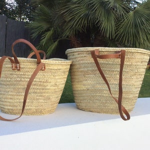 May include: Two woven straw tote bags with brown leather handles. The larger bag is on the right and the smaller bag is on the left. Both bags have a woven pattern.