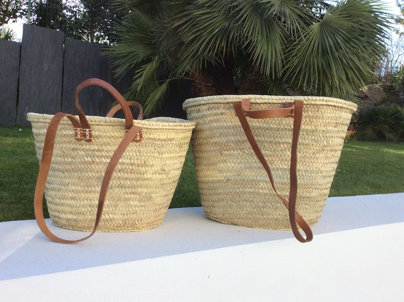 May include: Two woven straw tote bags with brown leather handles. The larger bag is on the right and the smaller bag is on the left. Both bags have a woven pattern.