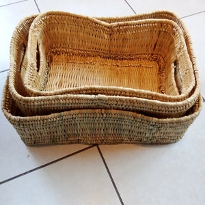May include: Three woven rectangular baskets of varying sizes nested together. The baskets are made of a light brown, natural fiber material. Each basket has a rectangular shape with rounded corners and built-in handles.