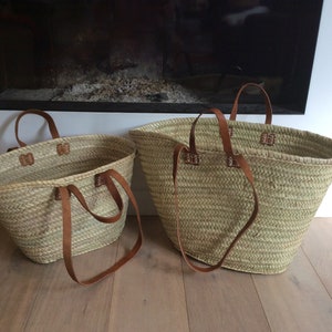 May include: Two woven straw tote bags with brown leather handles. The bags are different sizes and are sitting on a wooden floor.