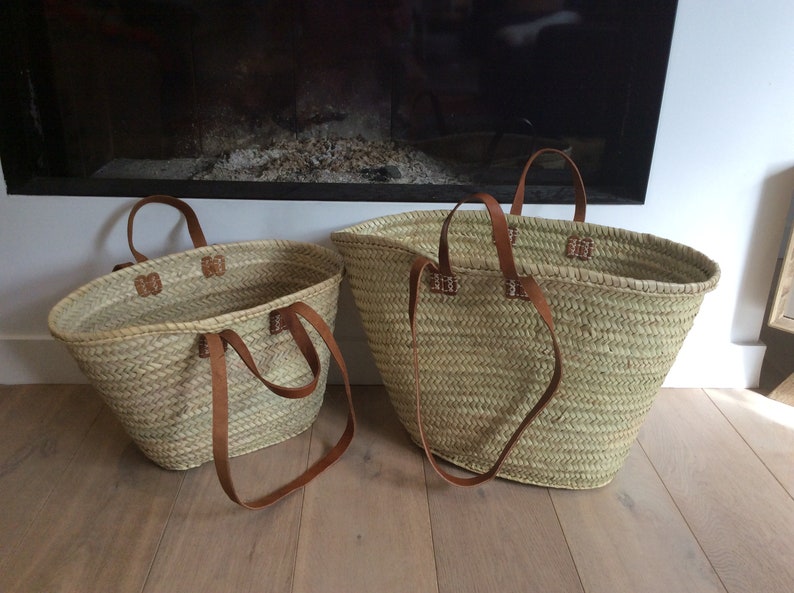 May include: Two woven straw tote bags with brown leather handles. The bags are different sizes and are sitting on a wooden floor.