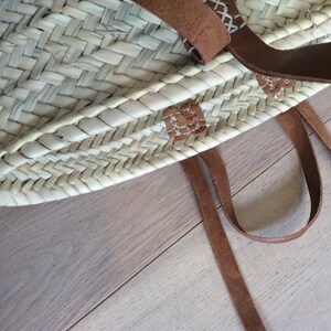May include: A woven straw tote bag with brown leather straps and stitching. The bag has a natural, light brown color and is made with a herringbone pattern.