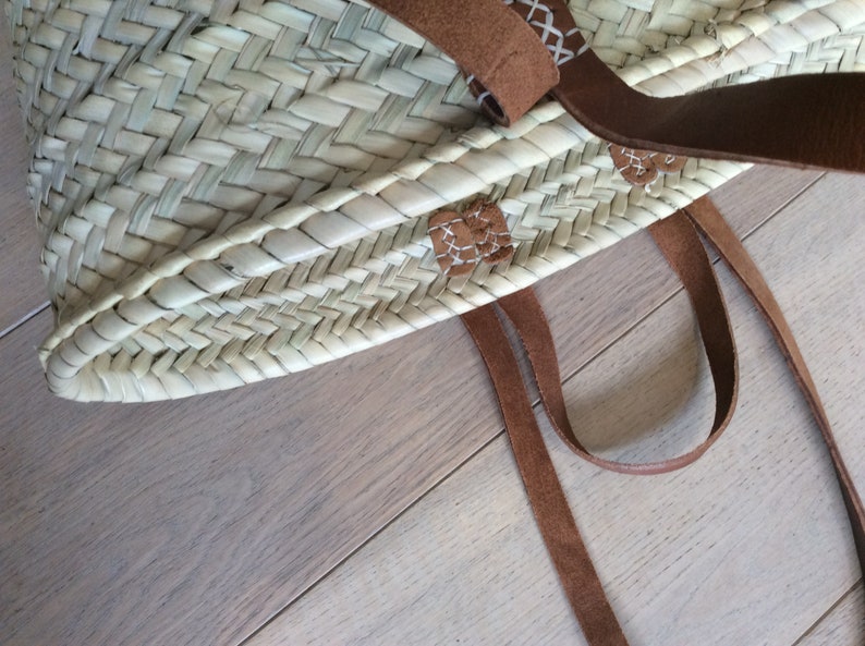 May include: A woven straw tote bag with brown leather straps and stitching. The bag has a natural, light brown color and is made with a herringbone pattern.
