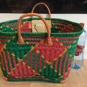 May include: A woven basket bag with green, red, and yellow stripes. The bag has leather handles and a small pocket on the inside. The bag is sitting on a wooden floor with a bottle of water next to it.