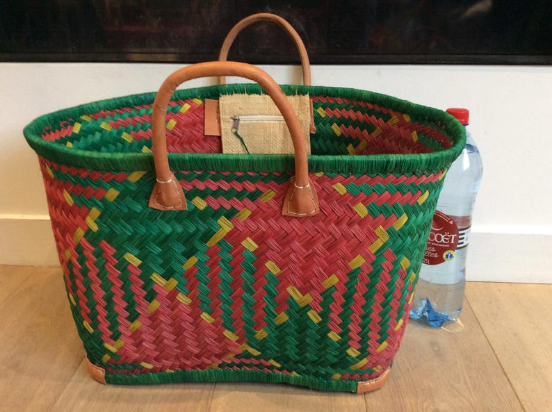 May include: A woven basket bag with green, red, and yellow stripes. The bag has leather handles and a small pocket on the inside. The bag is sitting on a wooden floor with a bottle of water next to it.