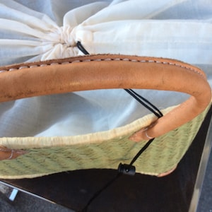 May include: A woven straw tote bag with a brown leather handle and a white cotton lining. The bag has a black drawstring closure.