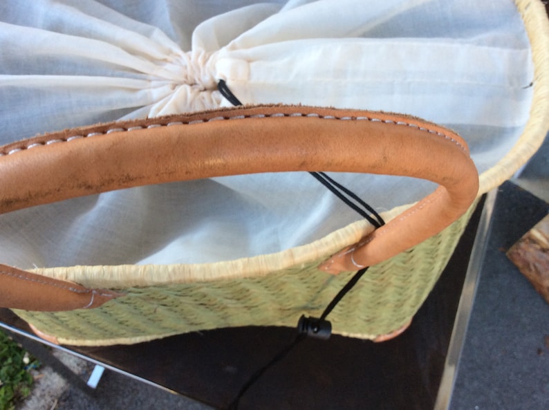 May include: A woven straw tote bag with a brown leather handle and a white cotton lining. The bag has a black drawstring closure.