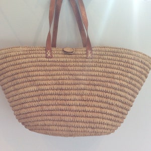 Puede incluir: Un bolso tote de paja tejida con asas y detalles de cuero marrón. El bolso tiene forma redondeada y cierre de botón. El bolso es de color beige natural y es adecuado para llevar objetos.