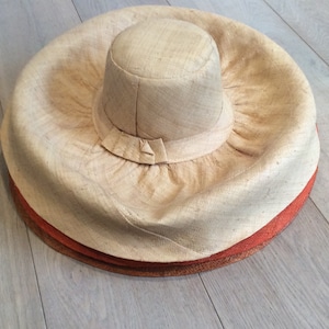 Puede incluir: Un sombrero de paja grande y de ala ancha con una corona redonda. El sombrero est&aacute; hecho de un material natural tejido y tiene un color marr&oacute;n claro. El ala del sombrero est&aacute; doblada hacia arriba y tiene un aspecto ligeramente arrugado.