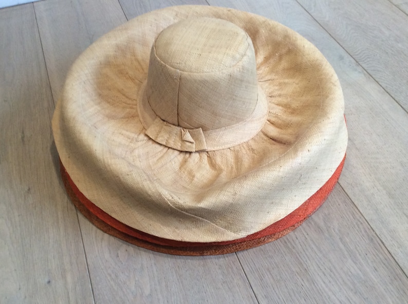 Puede incluir: Un sombrero de paja grande y de ala ancha con una corona redonda. El sombrero est&aacute; hecho de un material natural tejido y tiene un color marr&oacute;n claro. El ala del sombrero est&aacute; doblada hacia arriba y tiene un aspecto ligeramente arrugado.