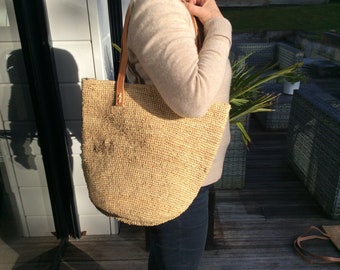 Könnte beinhalten: Eine große, hellbraune, gewebte Tragetasche mit Ledergriffen. Die Tasche ist aus einem natürlichen Material gefertigt und hat eine leicht abgerundete Form.