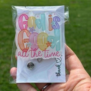 Könnte beinhalten: Ein Ausweis-Rollhalter mit dem Satz "God is Good all the time" in bunten Buchstaben im Blasenstil. Der Rollhalter befindet sich in einer transparenten Plastikhülle mit weißem Hintergrund und einer "Danke"-Nachricht.