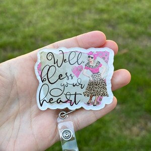 May include: A clear acrylic badge holder with a colorful design featuring a woman in a leopard print dress and the text "Well bless your heart". The badge holder has a clear plastic retractable badge reel.