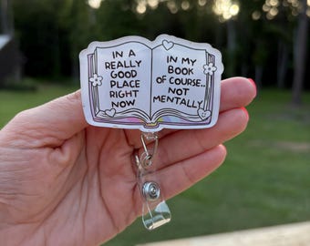 May include: A clear badge reel with a book-shaped design. The book has the text "IN A REALLY GOOD PLACE RIGHT NOW" on the left page and "IN MY BOOK OF COURSE.. NOT MENTALLY" on the right page. The design includes flowers and hearts.