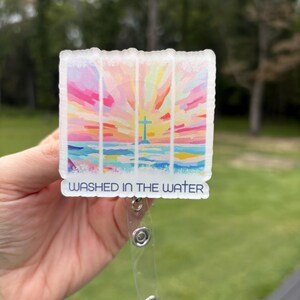 Puede incluir: Un portatarjetas con un diseño abstracto colorido que presenta una cruz y el texto "Washed in the Water". La obra de arte incluye tonos de rosa, amarillo, azul y naranja, que se asemejan a una puesta de sol sobre el agua. El carrete tiene una correa de plástico transparente y un clip de metal.
