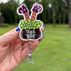 Pode incluir: Um porta-crachá com tema de Halloween com um caldeirão, bolhas verdes e pernas de bruxa. O caldeirão tem as palavras "Trick or Treat". As pernas da bruxa são roxas e pretas com meias listradas em laranja e roxo.