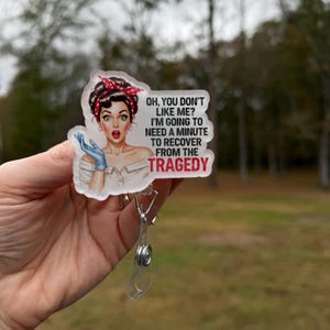 May include: A clear badge reel with a retro-style illustration of a woman with a red polka dot headband and the text "OH, YOU DON'T LIKE ME? I'M GOING TO NEED A MINUTE TO RECOVER FROM THE TRAGEDY."