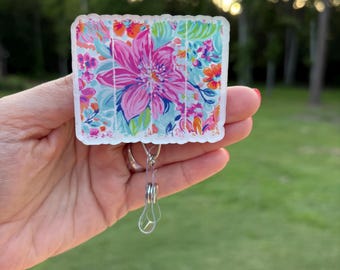 May include: A floral badge reel with a vibrant design featuring a large pink flower and colorful accents of blue, orange, and purple. The badge reel has a clear plastic clip and a retractable cord.