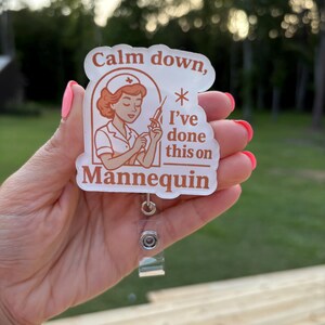 May include: A badge reel with a nurse illustration and the text "Calm down, I've done this on Mannequin." The design features a nurse in a vintage style uniform holding a syringe. The badge reel is held by a person with pink nail polish.