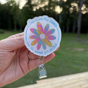 May include: A round badge reel with a floral design in shades of pink, yellow, and blue. The badge reel has the text "focus on the GOOD and the GOOD gets BETTER" in a handwritten style. The reel has a clear plastic clip and a retractable cord.
