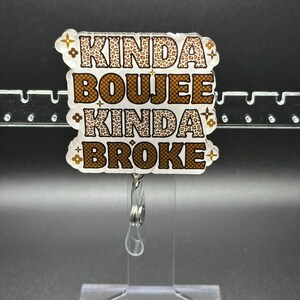 May include: A badge reel with the text "KINDA BOUJEE KINDA BROKE" in brown and leopard print lettering. The badge reel is clear and has a retractable cord and a clip on the back.