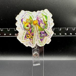 May include: A Mardi Gras-themed badge reel featuring a bow design with purple, green, and gold accents. The bow includes images of a mask, a crown, and a top hat. The words "king cake", "beads", and "crawfish" are written around the bow.