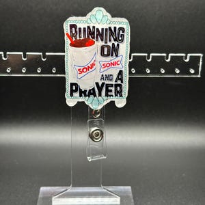 May include: A blue and white badge holder with a Sonic drink and the text "Running on Sonic and a Prayer".