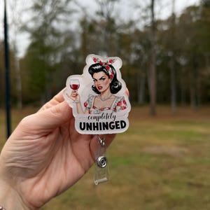 Könnte beinhalten: Ein durchsichtiges Ausweis-Jojo mit einer Retro-Illustration einer Frau, die ein Glas Rotwein hält. Die Frau hat dunkles Haar, roten Lippenstift und ein Blumenstirnband. Der Text "completely UNHINGED" steht darunter.