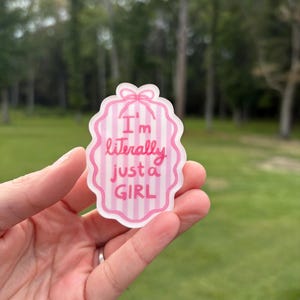 May include: A pink and white sticker with the text "I'm literally just a GIRL" in pink lettering. The sticker has a scalloped edge and a bow at the top. The background is a blurred outdoor scene.
