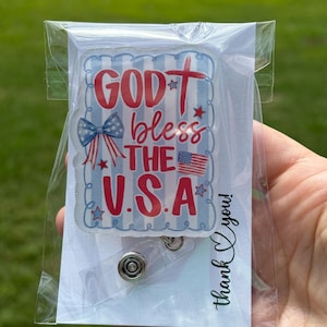 May include: A badge reel with a patriotic design. The badge reel features the text "GOD bless THE U.S.A." in red and white, along with a small American flag and a blue bow. The reel is packaged in a clear plastic sleeve.