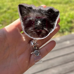 May include: A clear plastic badge holder with a black and white dog photo inside. The holder has a silver retractable clip.