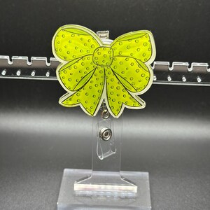 May include: A lime green bow-shaped badge reel with black polka dots. The badge reel has a clear plastic retractable cord and a metal clip. The bow design is outlined in black.