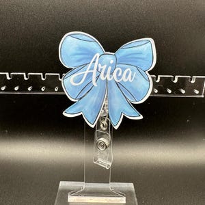 May include: A blue and white personalized badge holder with a bow and the name "Arica" printed on it.