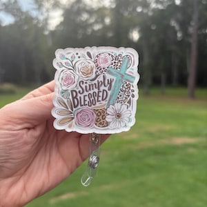 May include: A badge reel with a floral and leopard print design, featuring the words "Simply Blessed" and a turquoise cross. The badge reel has a clear plastic clip and a retractable cord.