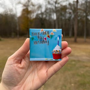 May include: A square magnet with a blue background and the text "Days Until My Birthday." The magnet features a cupcake with a lit candle and colorful sprinkles. The magnet is held in a hand.