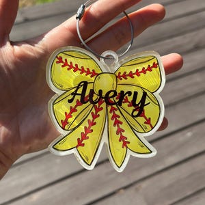 Może przedstawiać: Żółto-czerwona kokarda w kształcie piłki softball z napisem Avery w kolorze czarnym. Kokarda jest przymocowana do srebrnego metalowego pierścienia.